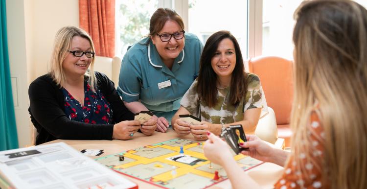 Three woman and a nurse sitting round a desk in a hospital day room playing a board game
